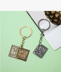 Two keychains shaped like open books with decorative elements on a green surface.